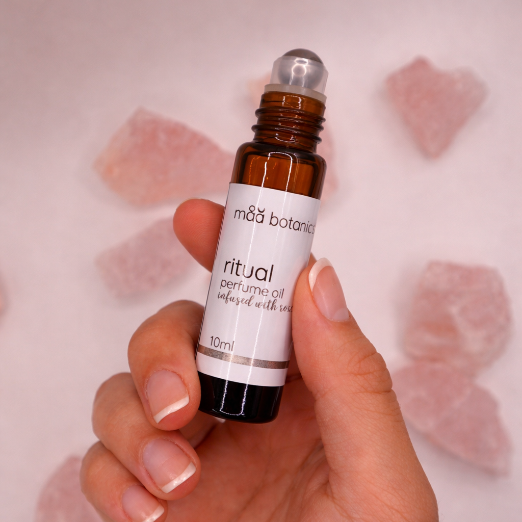 Hand holding a small brown rollerball bottle labeled 'ritual' against a soft pink background with rose quartz.