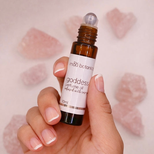 Hand holding a bottle of maa botanicals goddess perfume oil with a soft pink background.